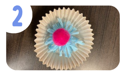 Labeled "two." A blue handmade pom pom in a yellow cupcake liner with a small pink pom pom on top.