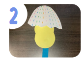 Labeled "two." A chick silhouette cut out of yellow paper wears a piece of white paper cut to look like the top of a cracked egg as a hat. A popsicle stick is attached to the bottom of the chick.
