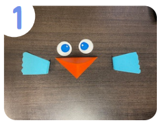 Labeled "one." On a brown table, two blue paper bird wings, an orange paper beak, and two white paper eyes are arranged.