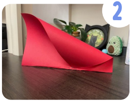 Labeled "two." A sheet of red cardstock that has been folded in half has one corner curled towards the center and the corner is stapled in place.