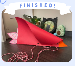 Labeled "Finished!" A photo of a completed red kite sitting on a desk that has been decorated to resemble a bird.