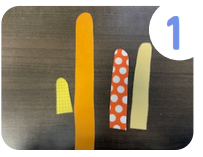 Labeled "one." Four strips of paper in various colors, patterns, and sizes that have been cut into strips with rounded tops. They sit on a brown table. 