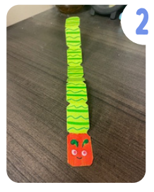 Labeled "two." A green and red paper caterpillar with a striped, segmented body, pink googly eyes, and a smiling face.