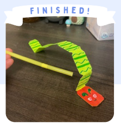Labeled "Finished!" A red and green paper caterpillar is folded into an arch on a wooden surface. A yellow straw is positioned behind the front of the arch.